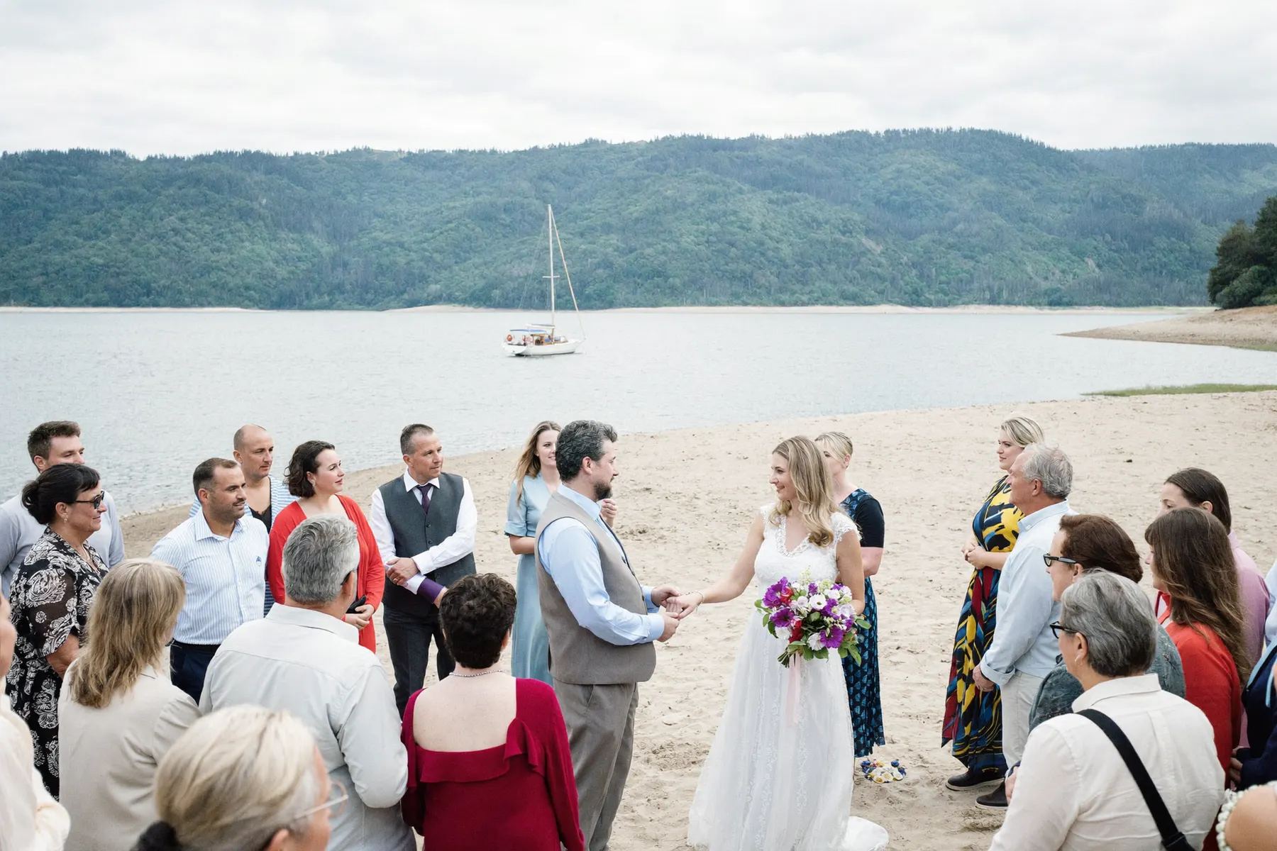 A beach ceremony at Deep Cove with guests standing and a sailboat on the water