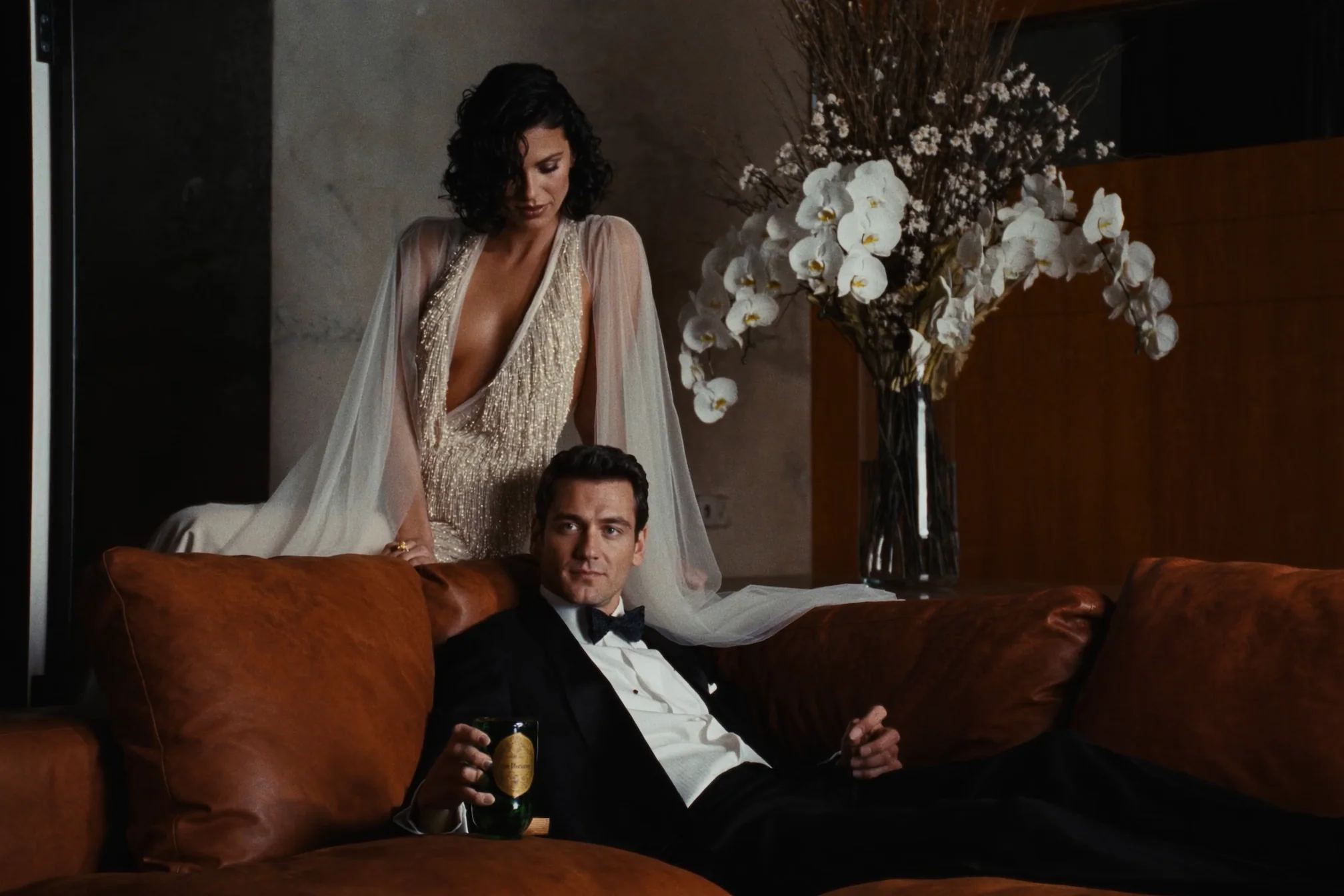 Bride in a beaded cape beside her groom on a leather sofa, white flowers behind them