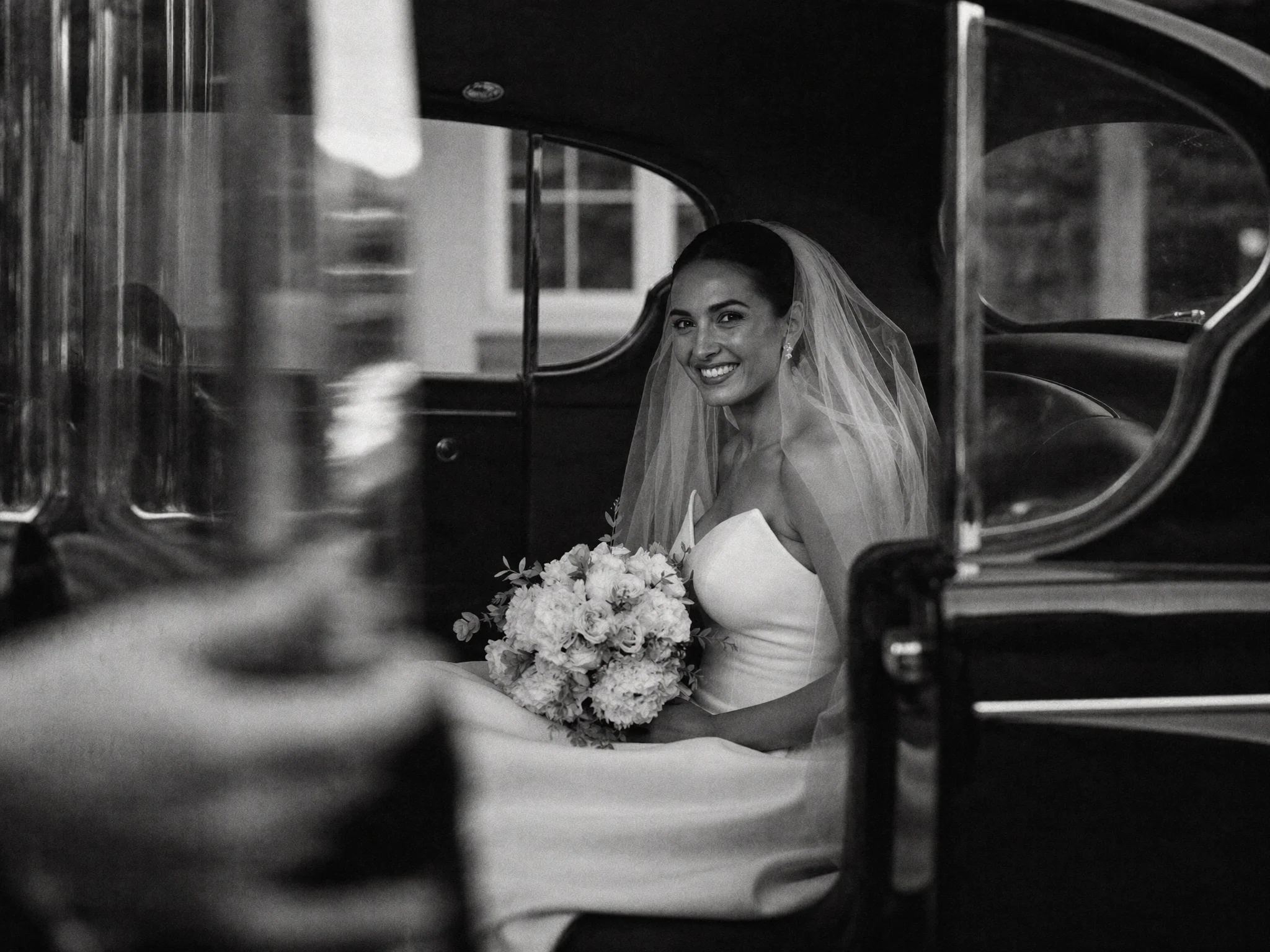 Bride holding her bouquet at the open door of a vintage car, shot through the window
