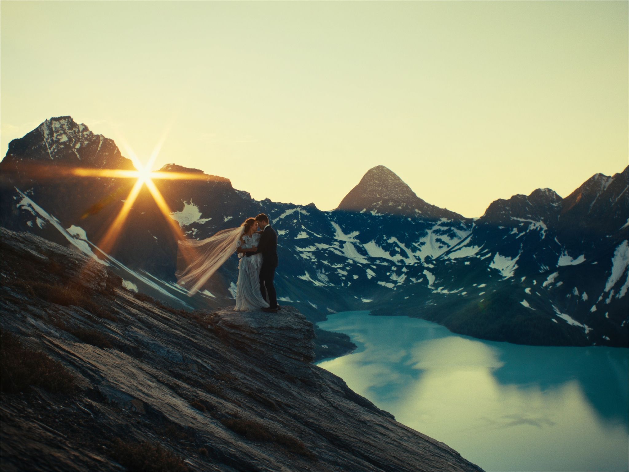 A couple on a high ridge with a veil catching the sunburst over a glacial lake in the Rockies