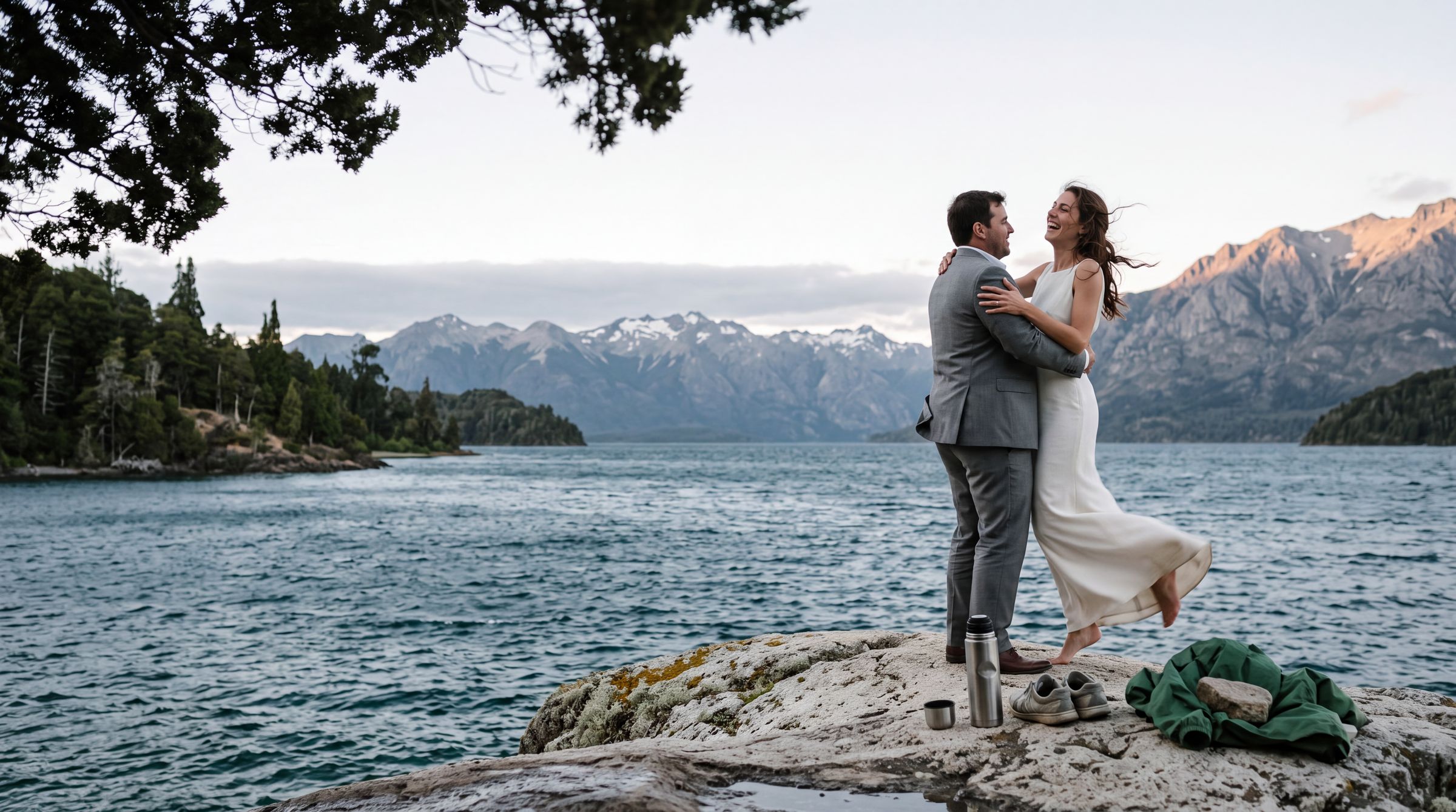 A couple on the rocks at the edge of Lago Nahuel Huapi below the Llao Llao peninsula, the Andes catching the last light behind them | Bariloche elopement photographer, Arman Arai