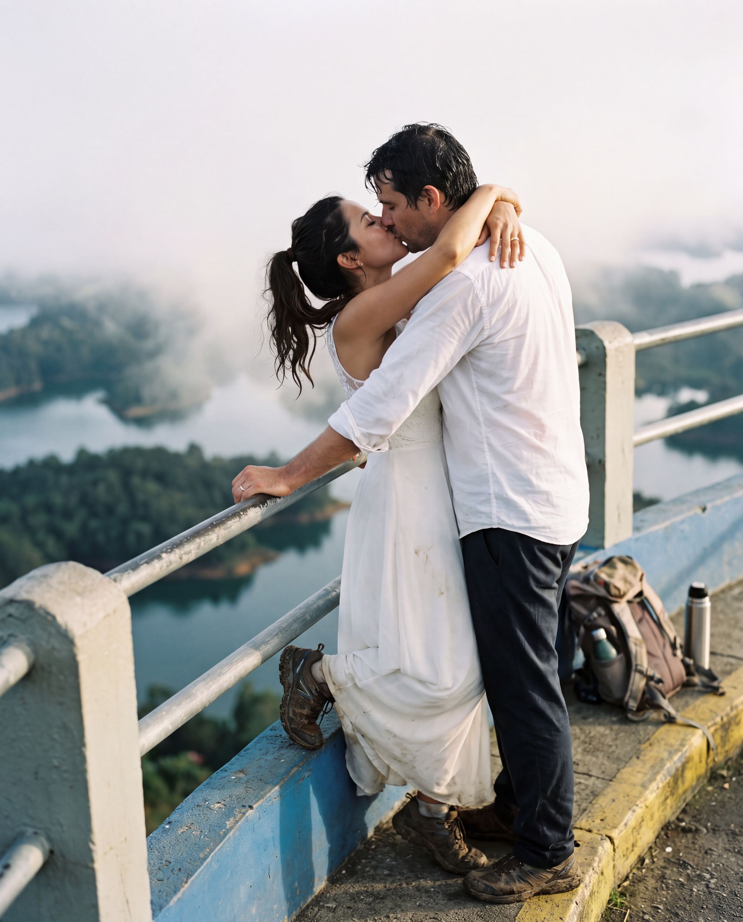 A bride in muddy trail shoes kissing her groom at the summit rail of La Piedra del Peñol at dawn, the island labyrinth of the Guatapé reservoir steaming two hundred metres below