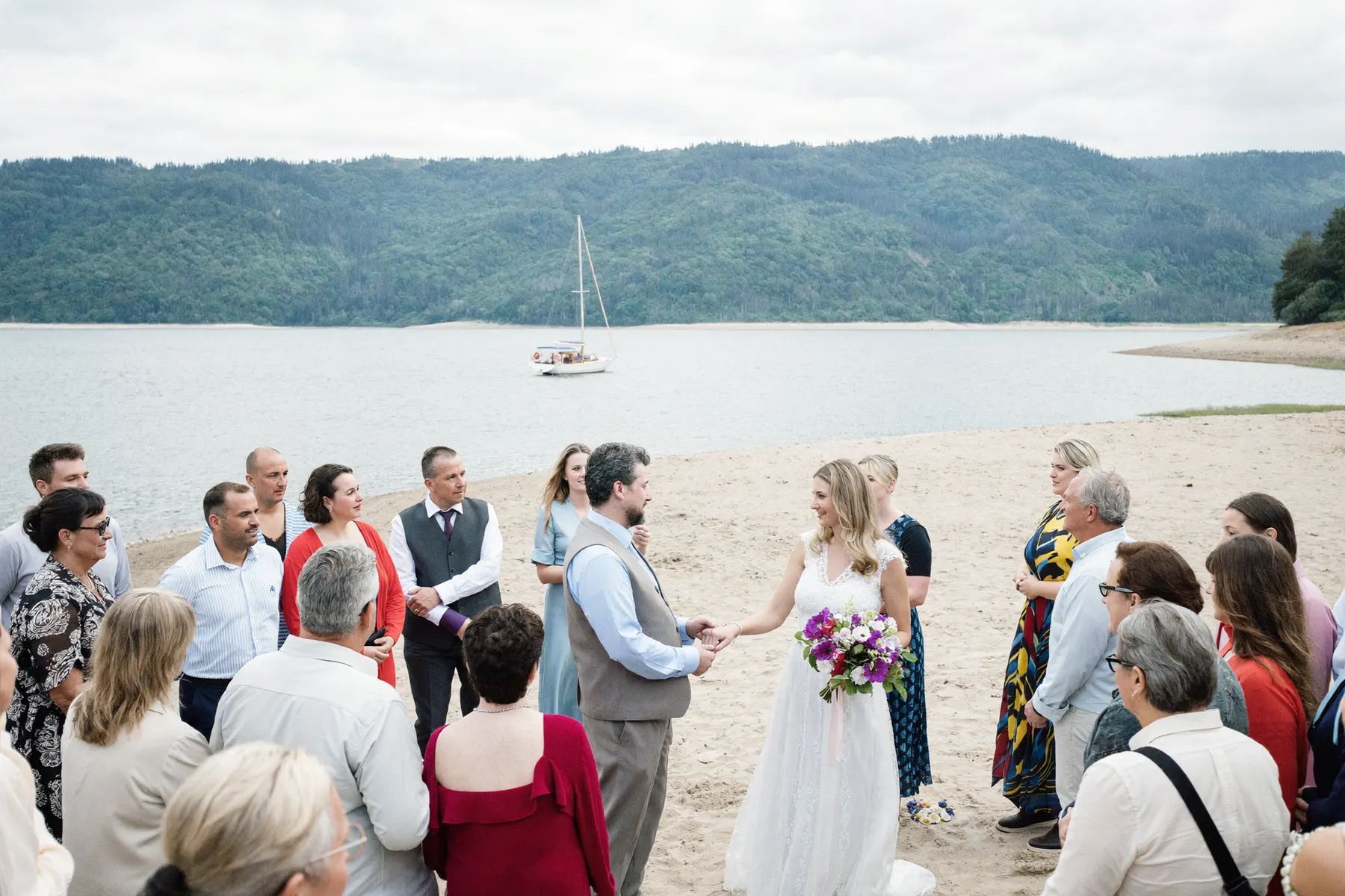 A beach ceremony at Deep Cove with guests standing and a sailboat on the water