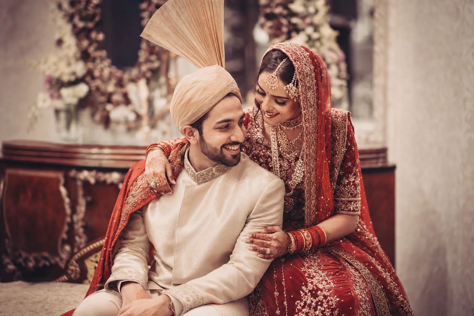 South Asian couple in a cream sherwani and a red and gold lehenga, seated close