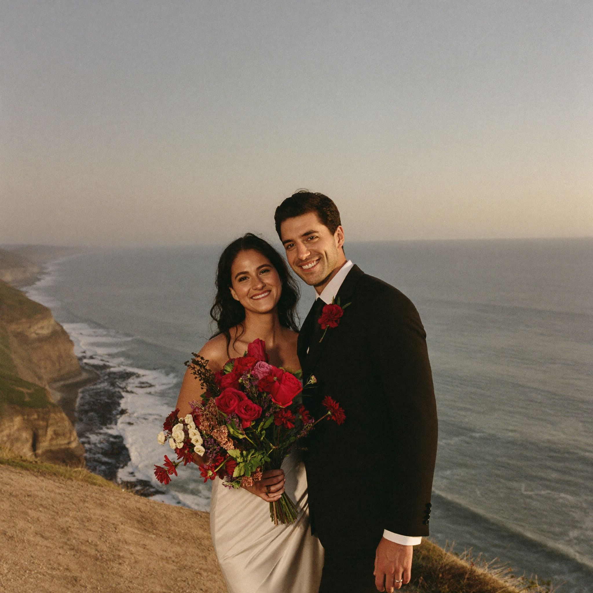 A couple on a clifftop above the Pacific holding a deep red bouquet