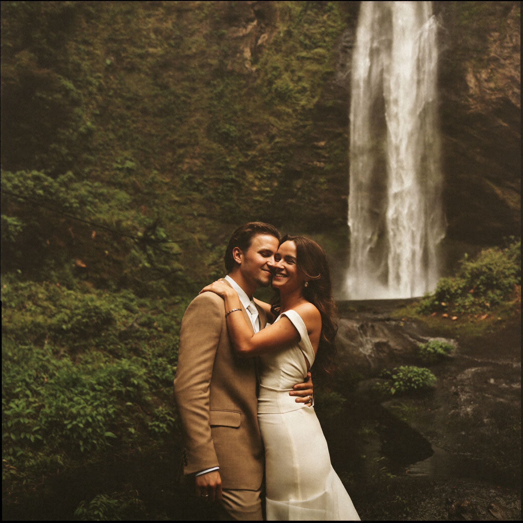 A couple standing soaked at the foot of a waterfall in the rainforest