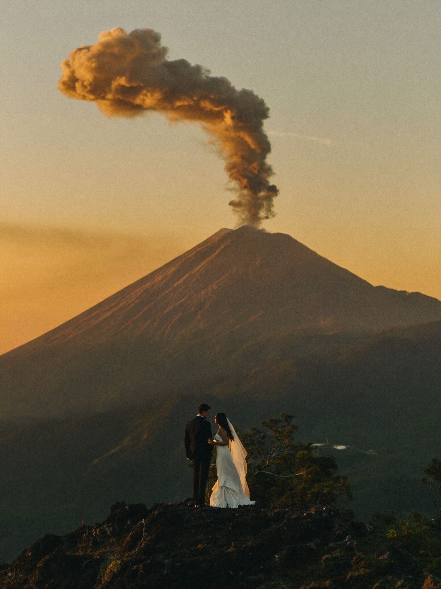 A couple standing on a ridge above the forest with the volcano cone behind them