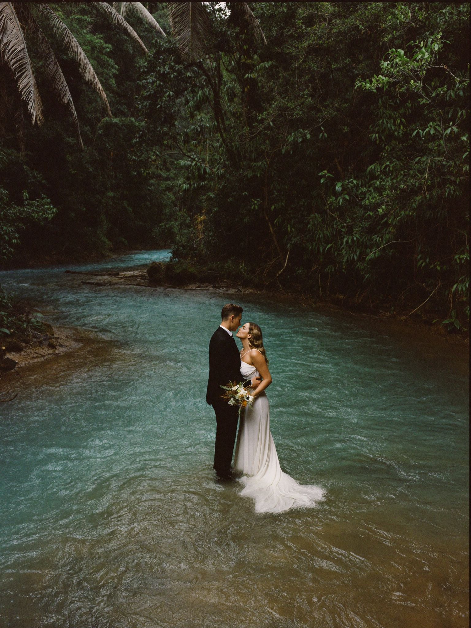 A couple standing in a turquoise river between rainforest banks