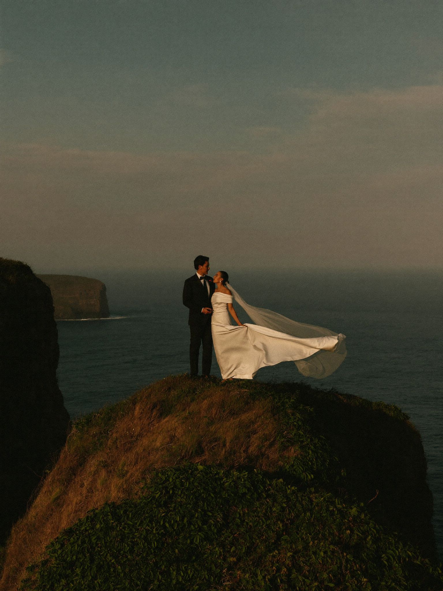 A couple on a green headland above a turquoise cove, her veil blowing out to the side