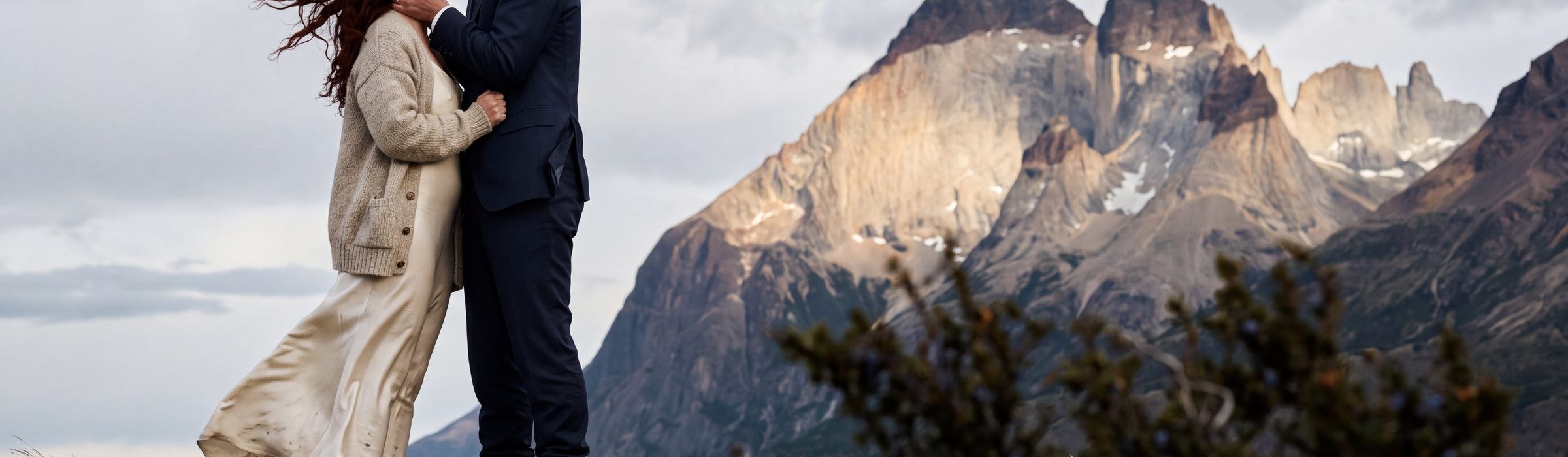 A couple on a wind-blasted moraine bluff in Torres del Paine, his hands on her face and a kiss to her forehead, one shaft of sun breaking onto the Cuernos del Paine behind them while they stand in cloud shadow | Arman Arai